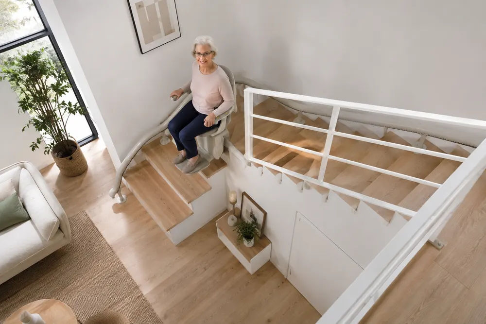 Vue plongeante d’un escalier tournant en bois clair équipé d’un monte-escalier. Le rail blanc part du bas près du séjour, longe la première volée, épouse le virage et se prolonge le long de la seconde volée, fixé sur les marches par une série de supports. Une femme âgée est assise sur le siège arrêté en haut de la première volée. L’escalier reste dégagé sur toute sa largeur et aucun percement n’a été fait dans le plancher ni dans les murs.